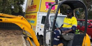 Construction worker operating a compact excavator near parked vehicles and a promotional signage truck, performing roadside work.