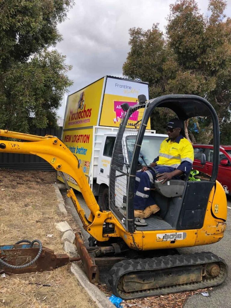 Construction worker operating a compact excavator near parked vehicles and a promotional signage truck, performing roadside work.